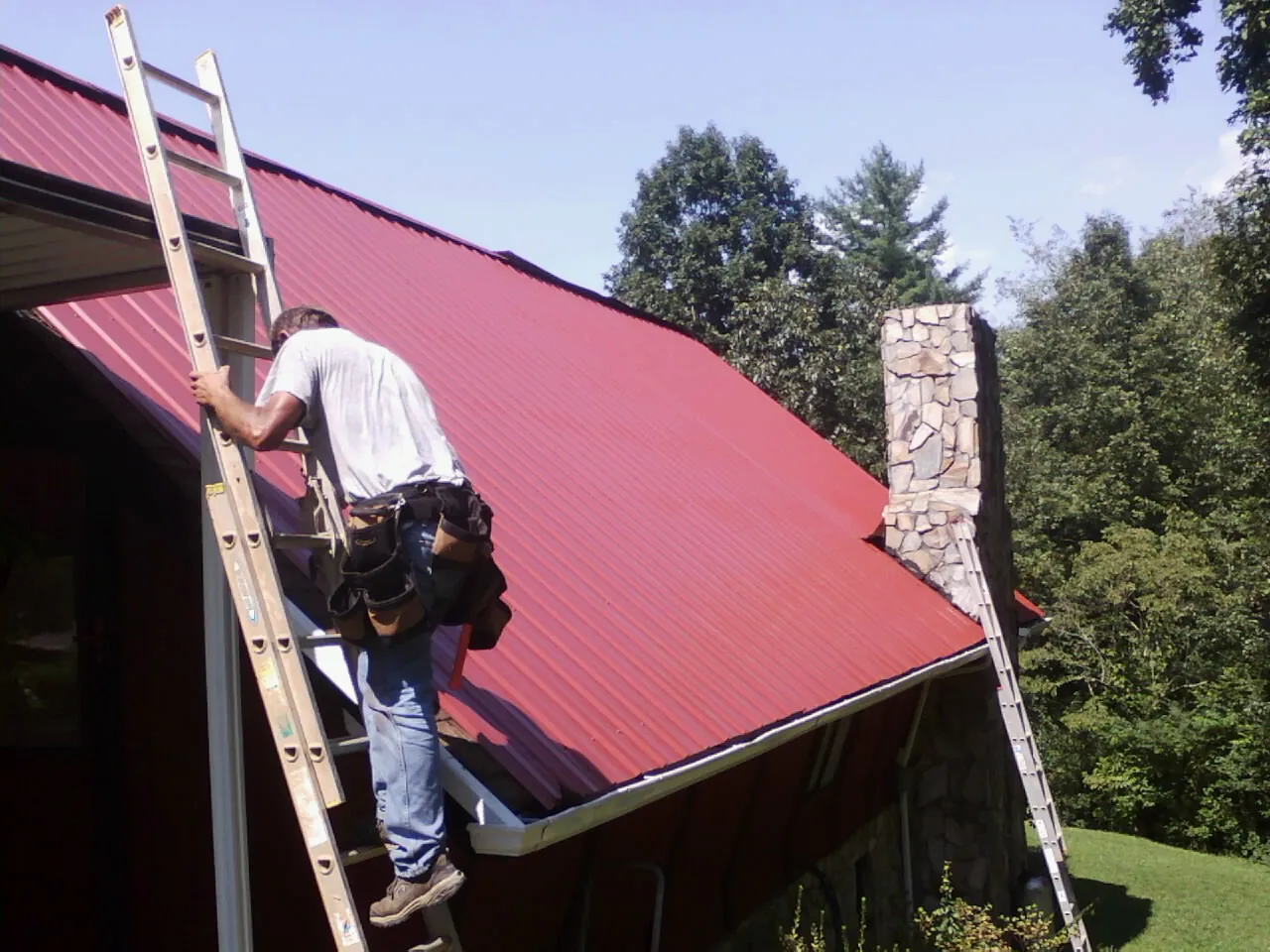Roofer on a ladder beside a red metal roof and stone chimney — Soffit & Fascia Repair by Roofing Brothers in Keyport